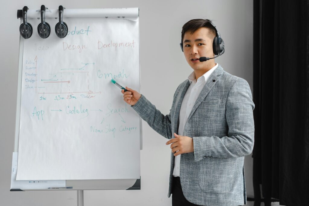 pexels-photo-7681136-7681136 Asian man in a gray suit giving a presentation with a flipchart, wearing a headset.